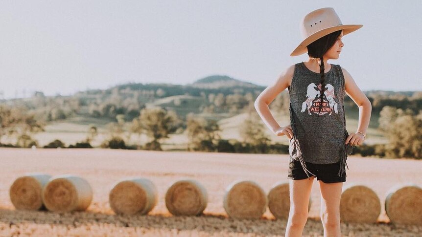 A young girl stands on top of a haybale while wearing her cowboy boots.