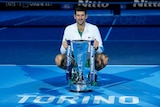 Novak Djokovic smiles as he sits on his haunches on the court, with a big trophy in front of him.