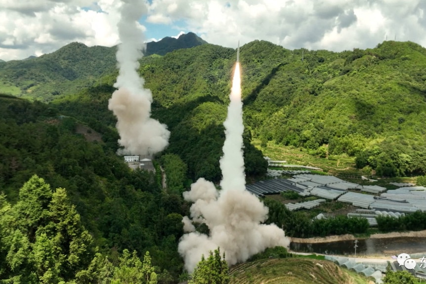 Missiles leave the ground in a cloud of smoke from between lush mountians.