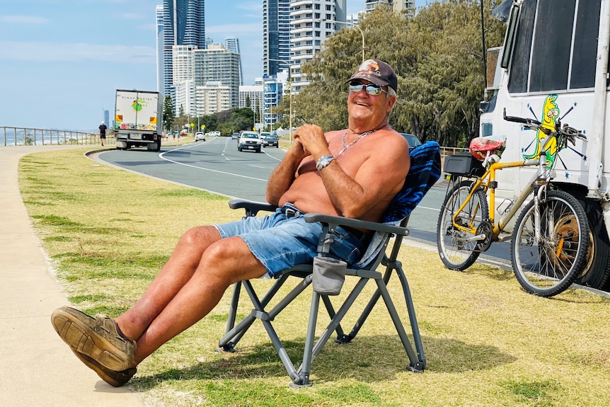Shirtless man sitting in camp chair in front of a bus and pushbike.