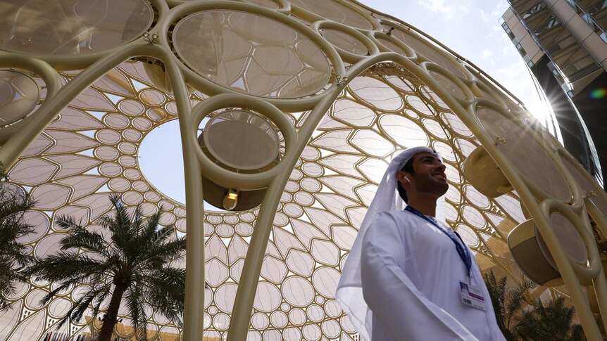 A man dressed in a traditional white Dishdasha walks underneath a towering yellow dome.