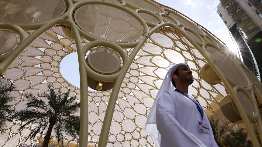 A man dressed in a traditional white Dishdasha walks underneath a towering yellow dome.