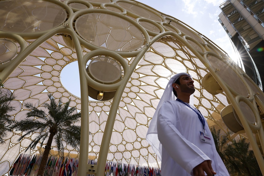 A man dressed in a traditional white Dishdasha walks underneath a towering yellow dome.