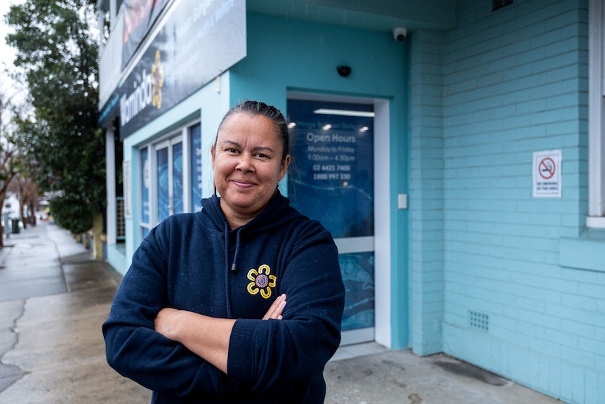 An Aboriginal woman stands with her arms crossed outside of a health clinic.