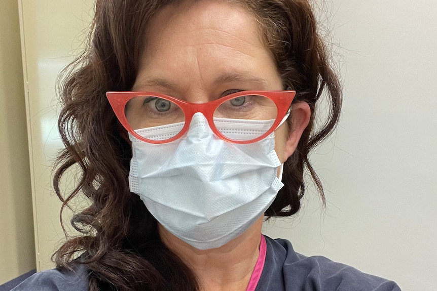 Annabel Crabb, a woman with auburn hair and red rimmed spectacles, takes a selfie in a surgical mask and navy medical gown
