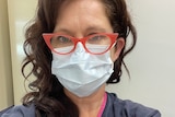 Annabel Crabb, a woman with auburn hair and red rimmed spectacles, takes a selfie in a surgical mask and navy medical gown