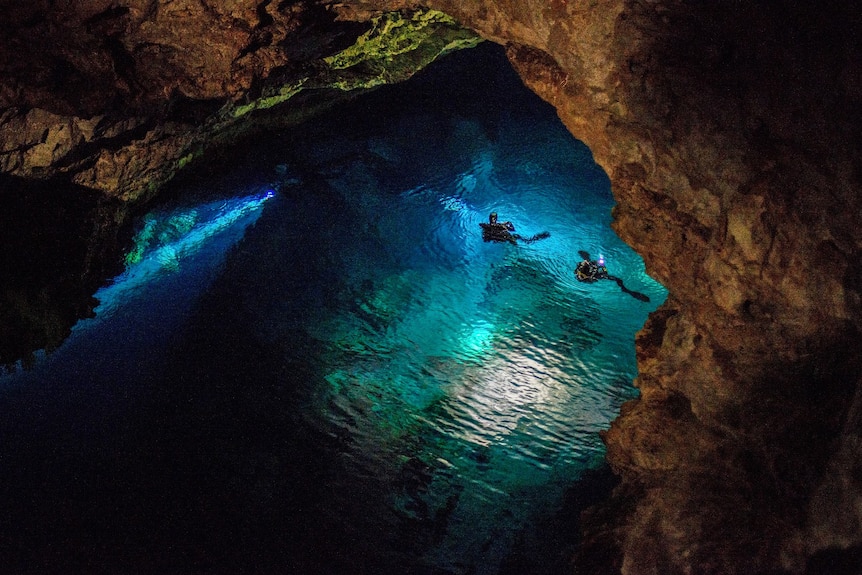 Two divers in a large body of clear blue water illuminated by a torch light.