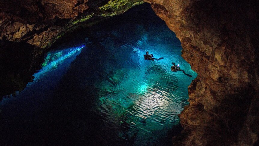 Two divers in a large body of clear blue water illuminated by a torch light.