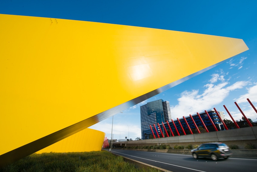 A large yellow beam juts out over a freeway, red poles in background