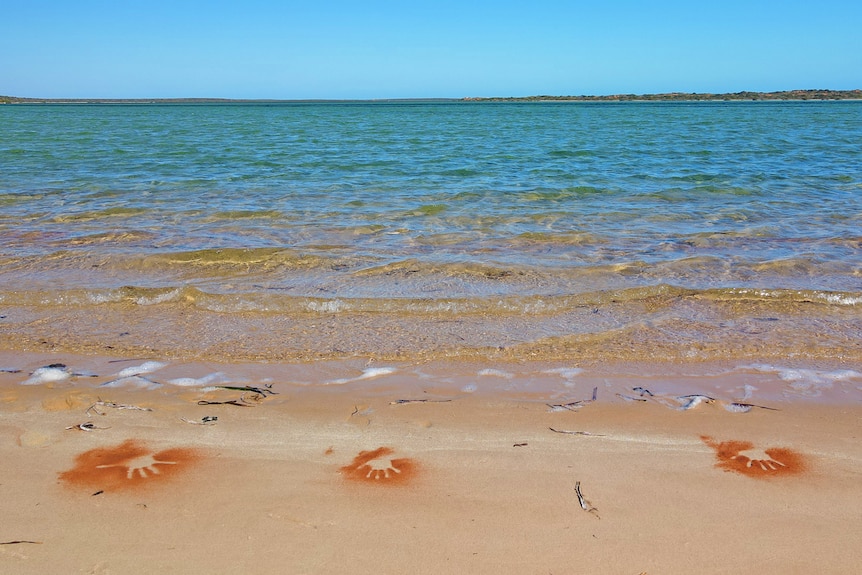 Outline of hands in red on white beach sand with ocean behind 