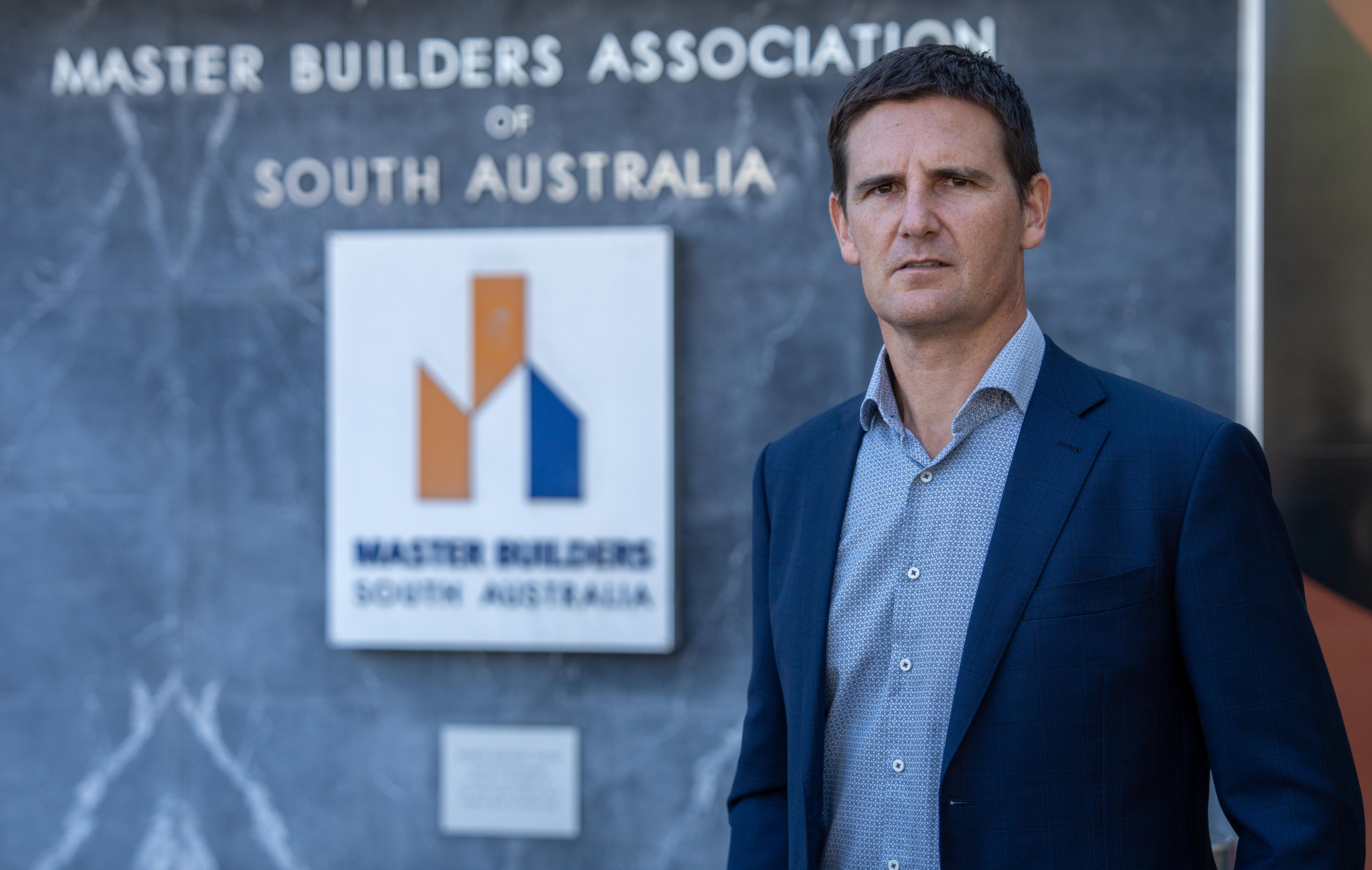 A an in a suit stands near a sign which reads Master Builders South Australia