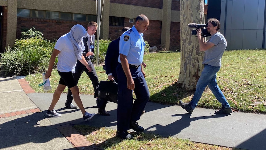 A man with a jumper obscuring his face walks across a lawn in front of a court house while being filmed by a TV cameraman.