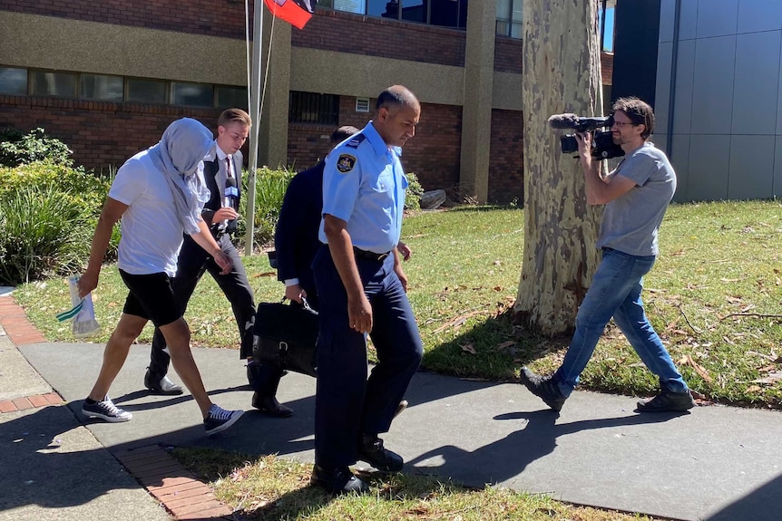 A man with a jumper obscuring his face walks across a lawn in front of a court house while being filmed by a TV cameraman.
