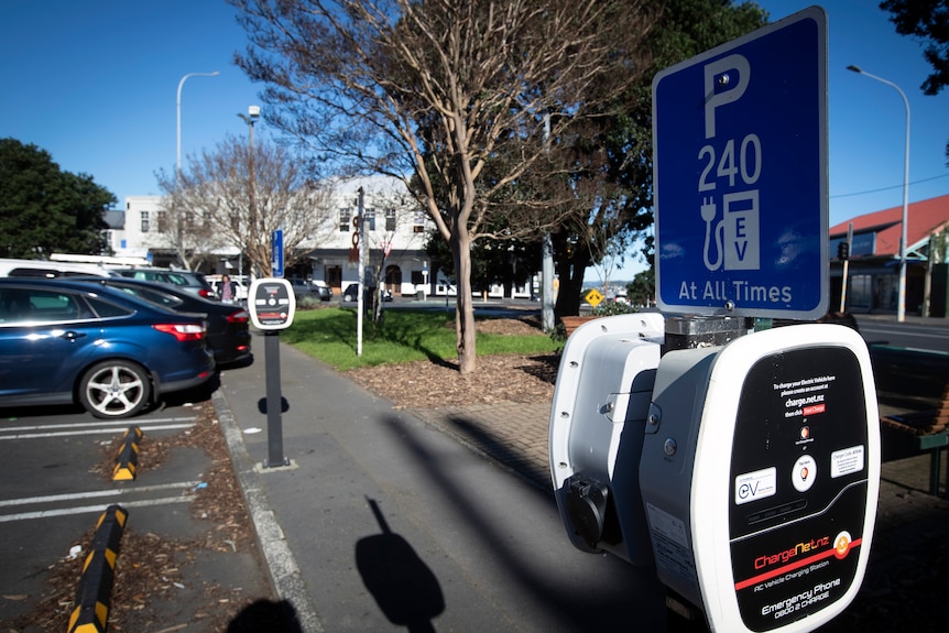 Close up of an electric vehicle charging station on a street with cars parked in background an trees on medium strip