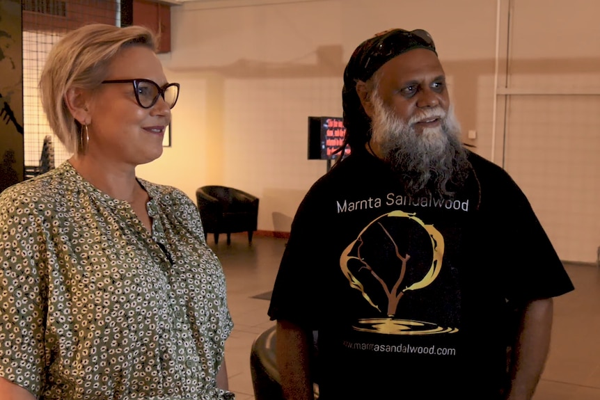 A woman with short blonde hair and glasses stands beside an older Aboriginal man in a large room.