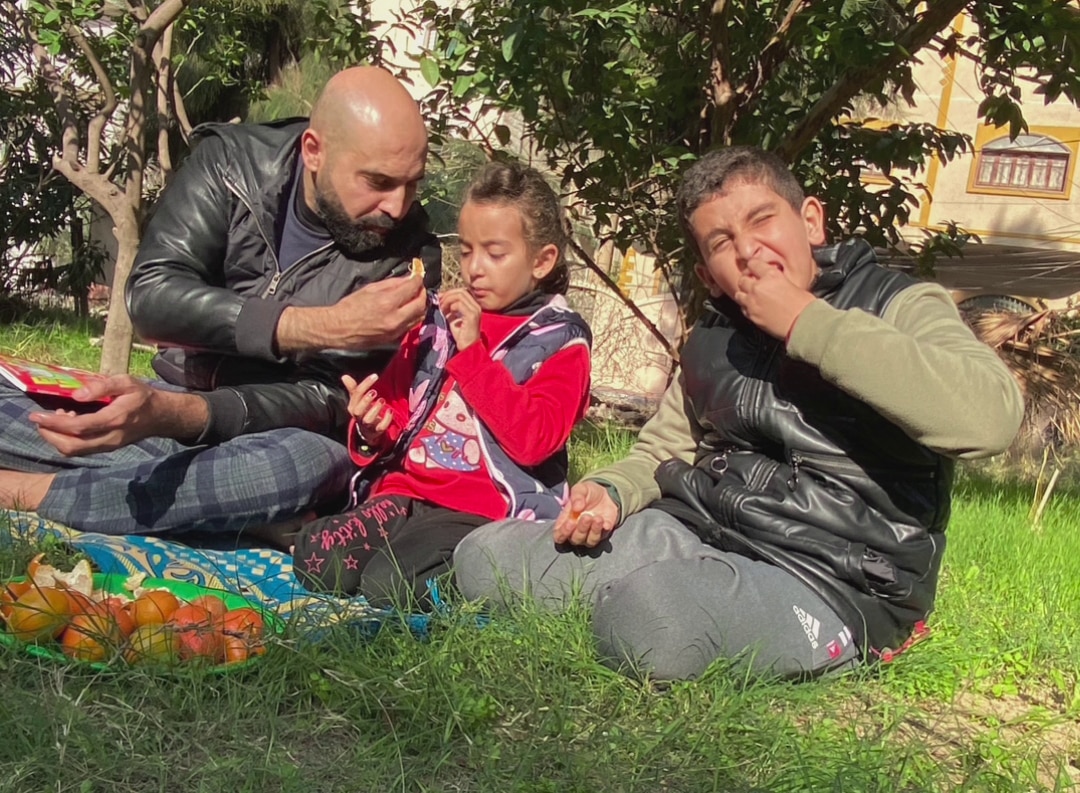 A father sits on the grass with his daughter and son eating food. 