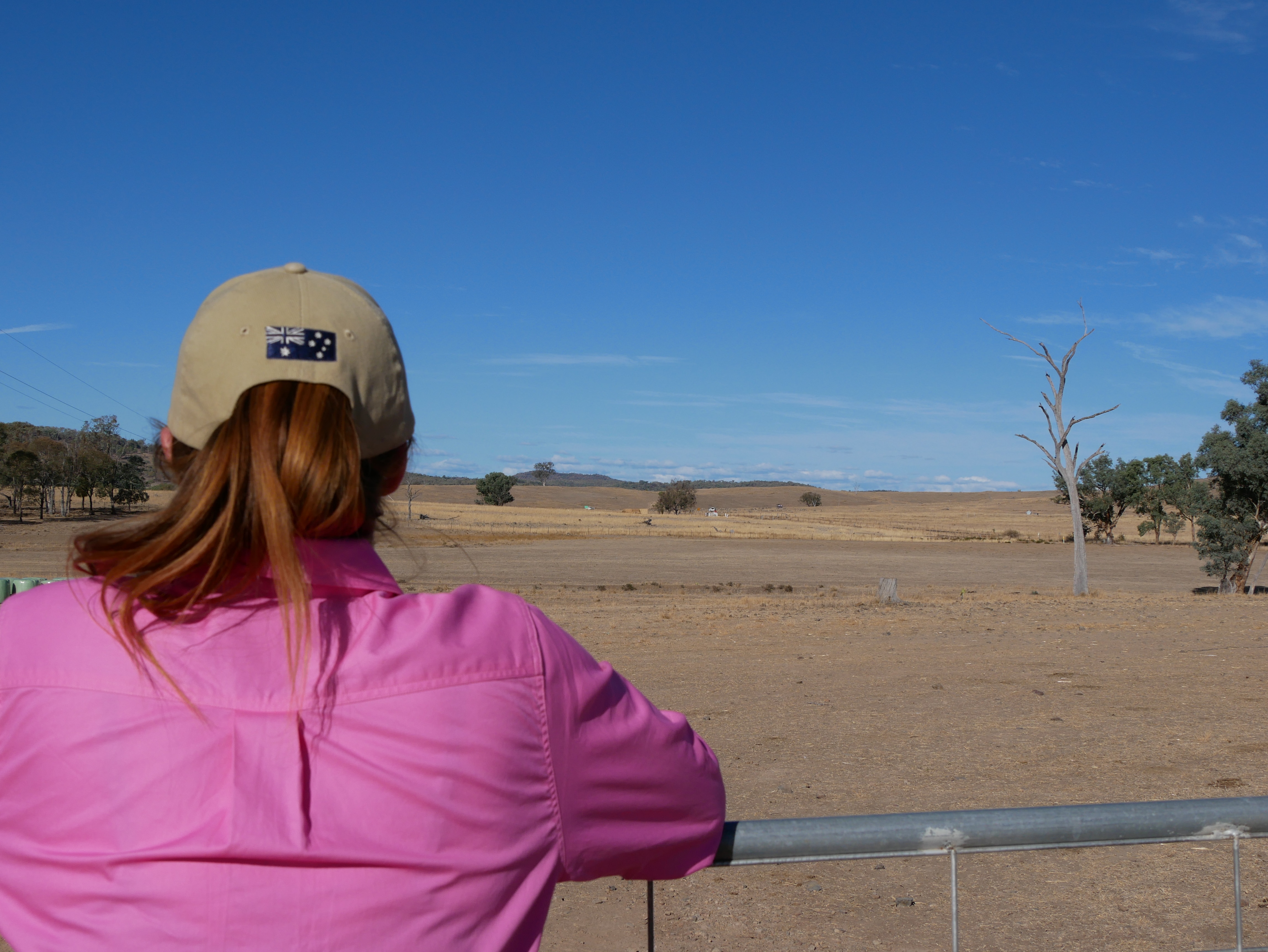 A woman in a pink shirt and cap looking out over a brown paddock.
