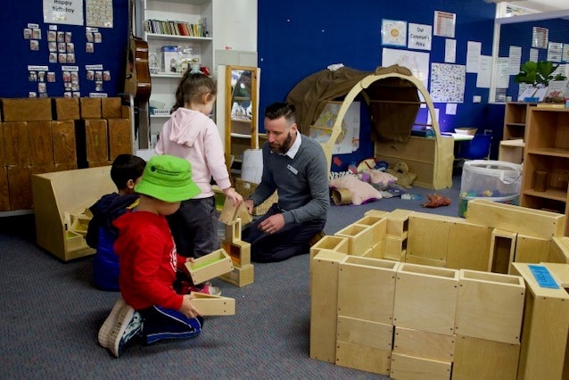 A teacher kneels on the floor facing his students as they show him their work