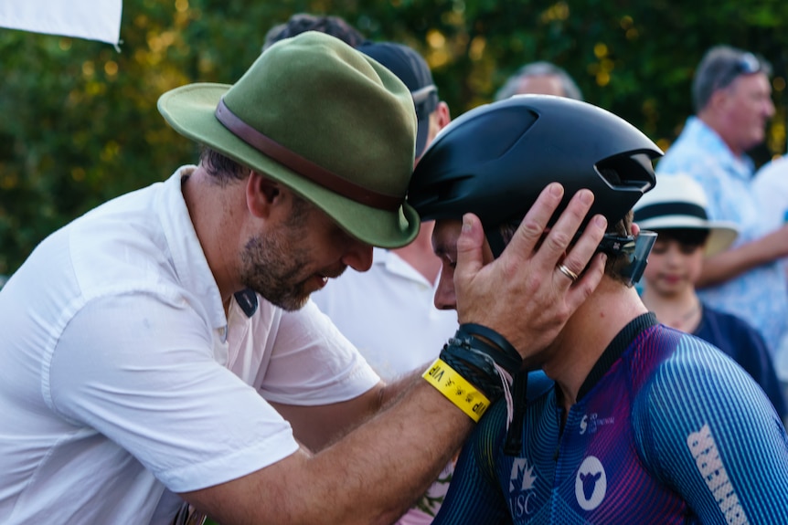 Two people touch foreheads with one person wearing a bike helmet, and the other a hat, white t-shirt.