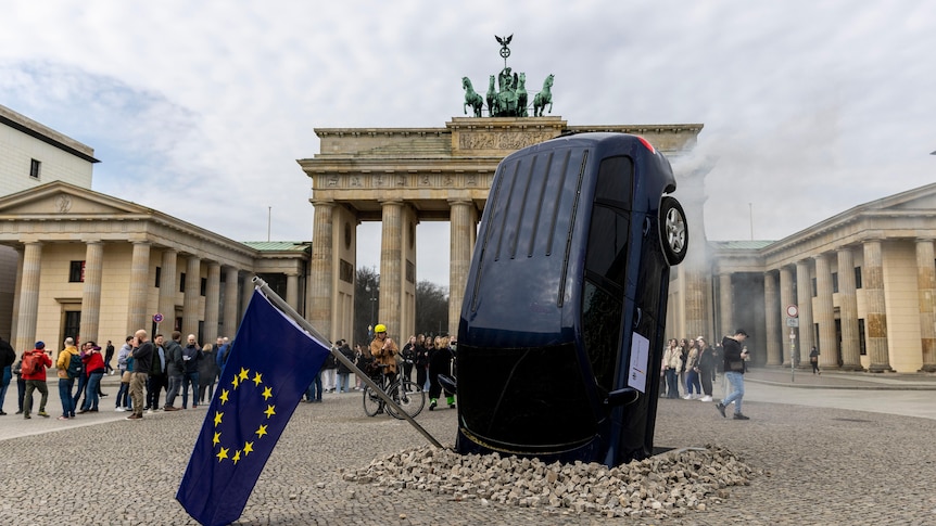 A black car stands on its end, with its bonnet buried in the cobblestones of a large open plaza in Berlin. 