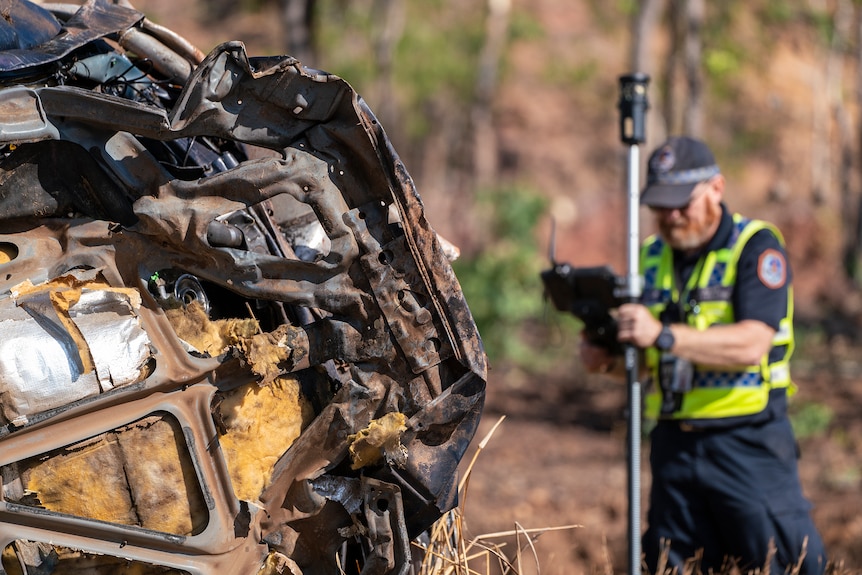 A police officer is holding equipment near a severely damaged car on its side. 