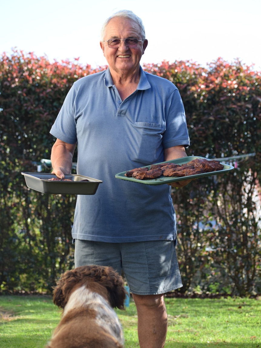 a man holding two trays of cooked mutton bird with a brown and white dog looking up a him