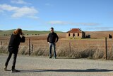 A woman takes a photo of a man posing in front of a tidy cottage in an empty field