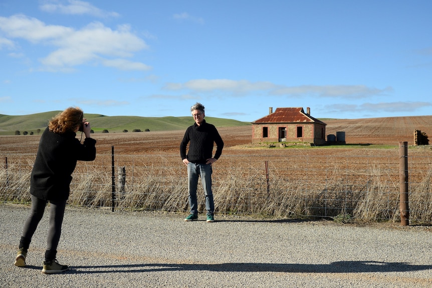 A woman takes a photo of a man posing in front of a tidy cottage in an empty field