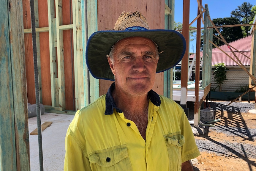 Builder with yellow high visual shirt and broad-brimmed hat stands in shade of tiny house frame under construction