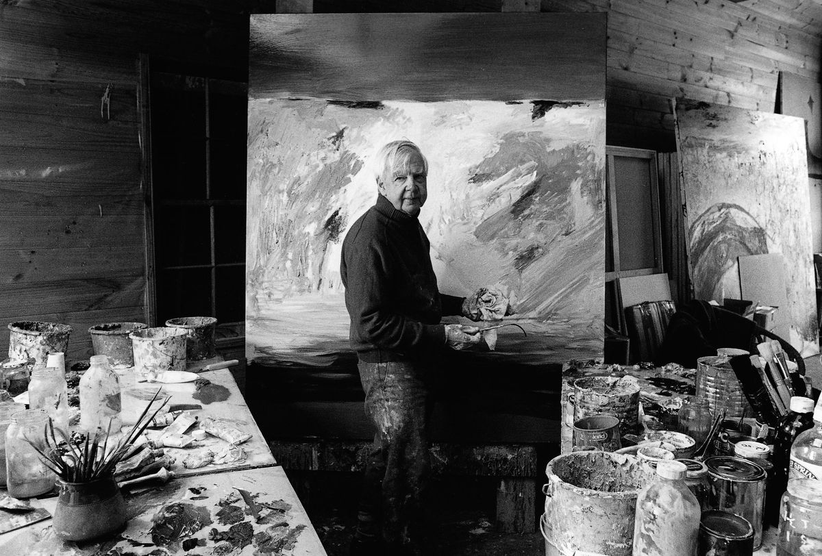 A black and white image of an older man with white hair standing in a painting studio.