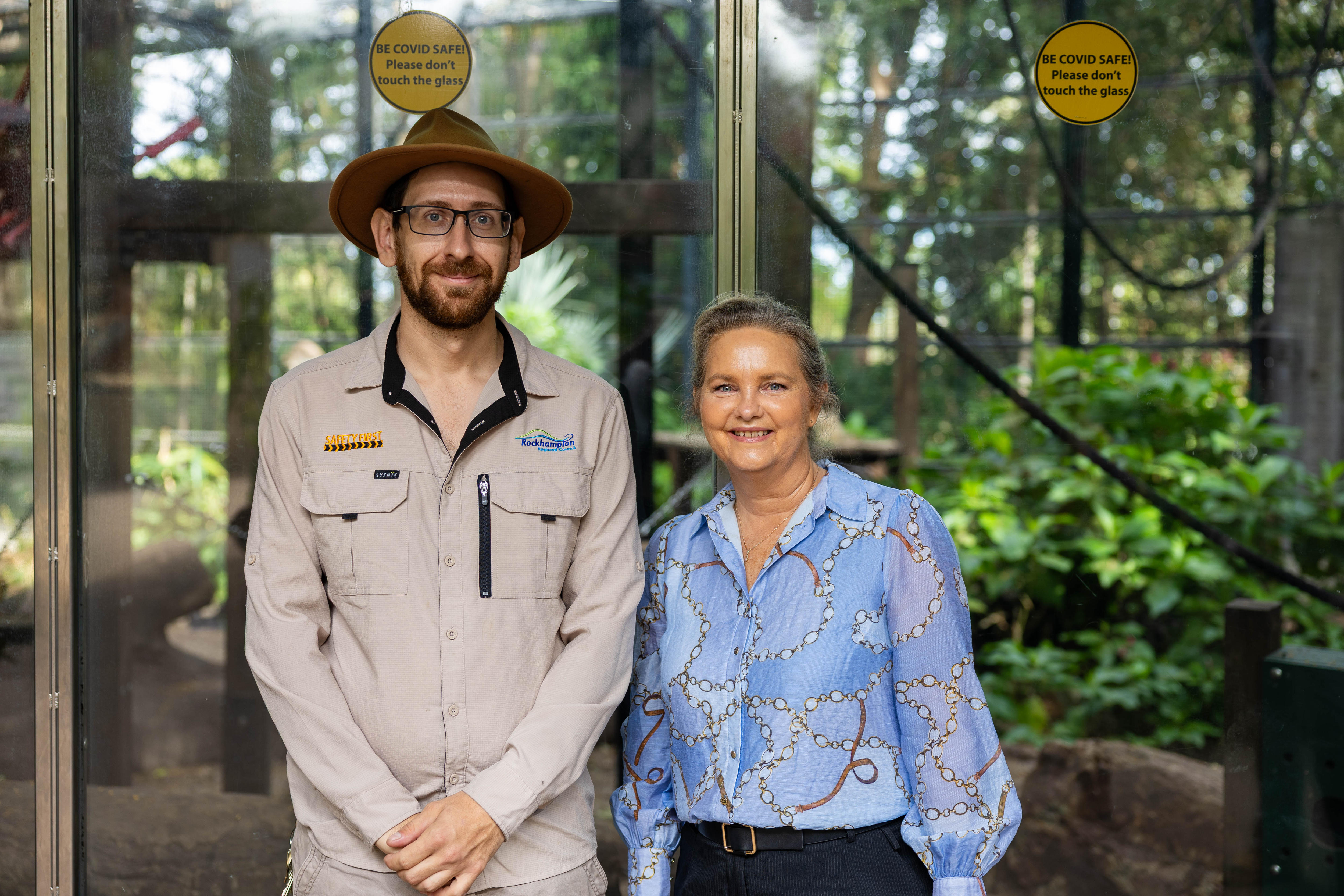 A man wearing a beige long sleeve collared shirt and hat, and a woman in a blue blouse smile in front of a chimp zoo enclosure.
