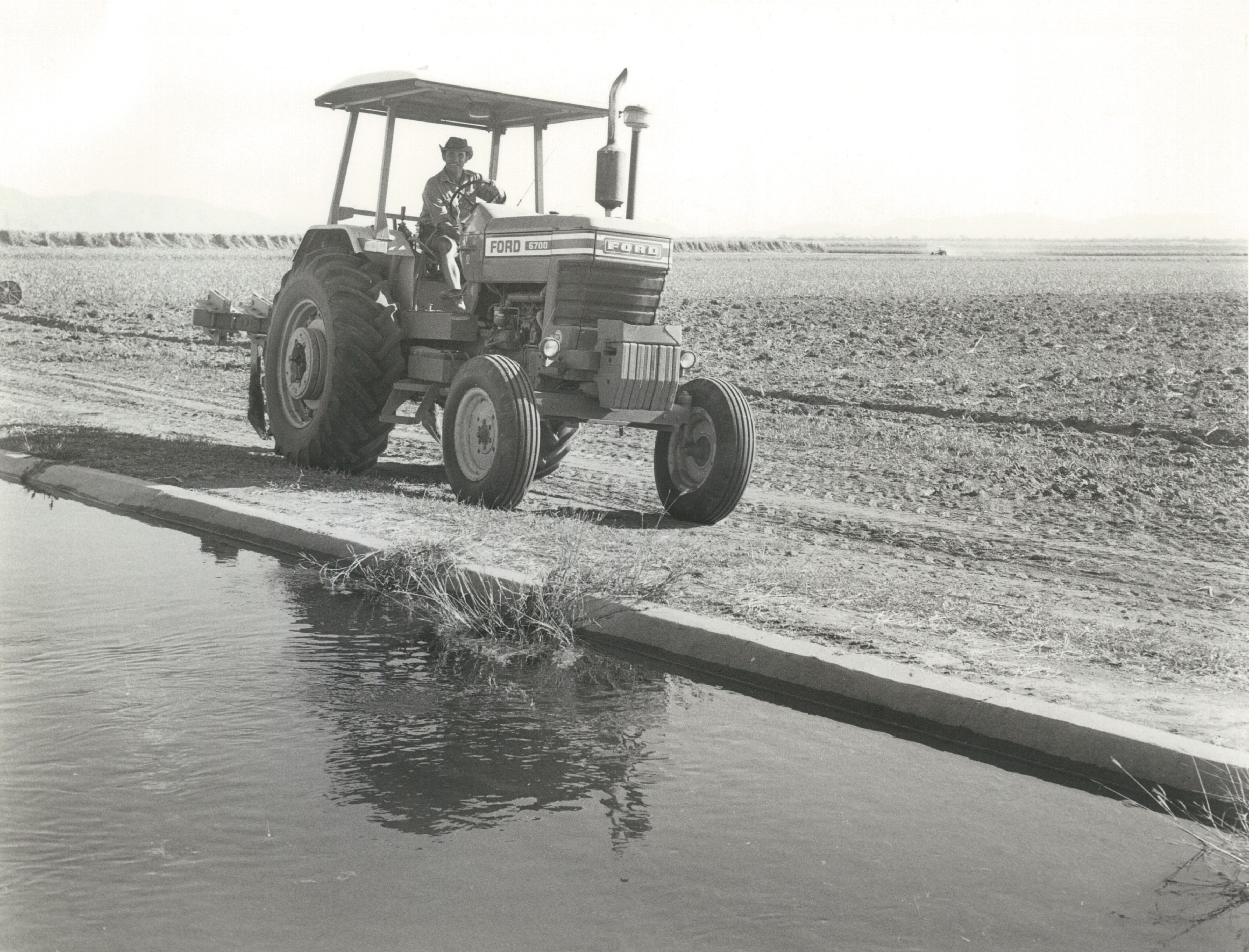 Uma fotografia em preto e branco de um homem dirigindo um trator próximo a um canal de irrigação em uma fazenda.