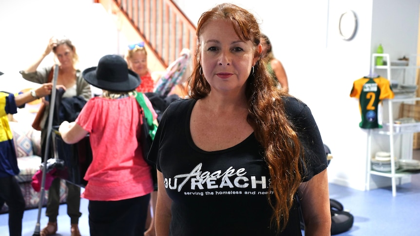 A woman with wavy red hair in an Agape Outreach t-shirt looks at the camera. Behind her people gather around tubs of clothes.