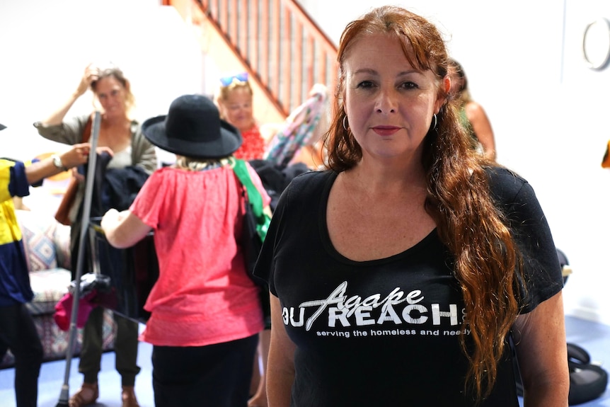 A woman with wavy red hair in an Agape Outreach t-shirt looks at the camera. Behind her people gather around tubs of clothes.