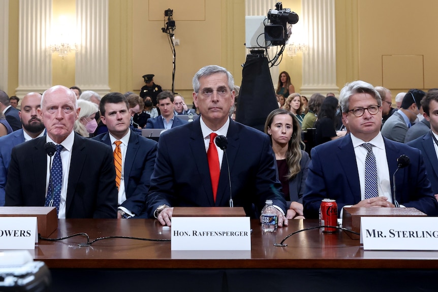 Three men in suits sit side by side looking stern during a January 6 committee hearing