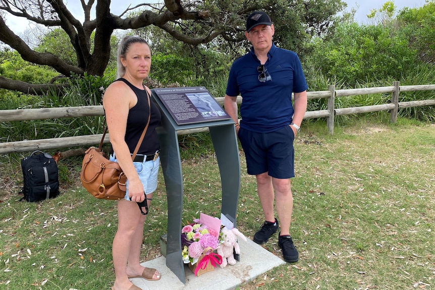 A man and a woman stand near a memorial plaque with flowers