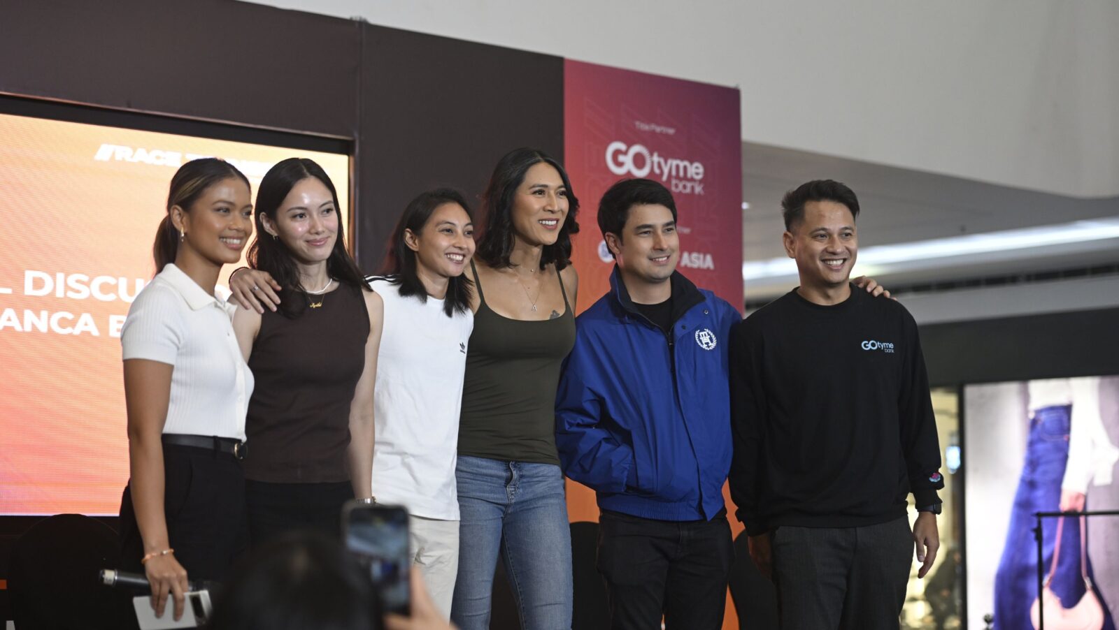 Four women and two men stand in a line on a stage with their arms around each other.