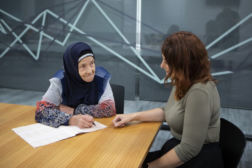 An elderly woman wearing a headscarf sits with a younger woman completing forms