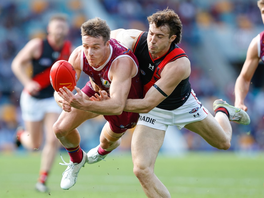 A Brisbane Lions player tries to handball the ball away in mid-air as he is tackled by an Essendon player.  