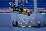 A man in a yellow shirt is mid-jump on a BMX bike. The Olympic rings are displayed on a wall in the background. 