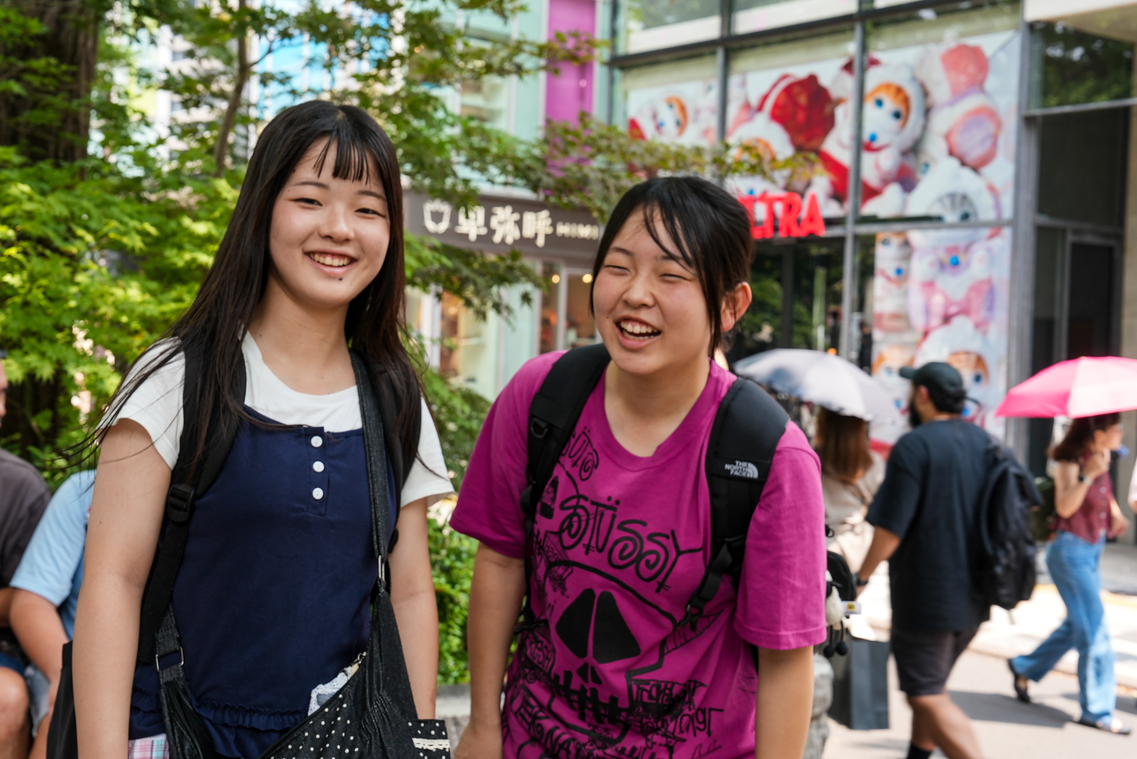 Two teenage girls smiling and laughing, while pedestrians carrying sun parasols stroll past on the street behind them.