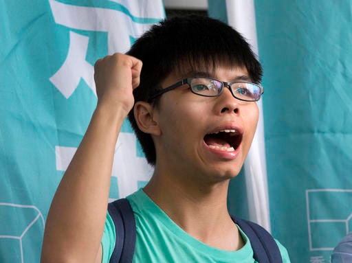 Hong Kong student activist Joshua Wong shouts with his hand raised in the air in a fist, wearing glasses and a backpack.