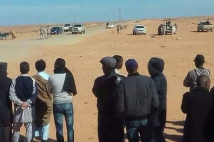 Families stand in the desert looking across at militiamen holding guns and weapons in their direction