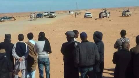 Families stand in the desert looking across at militiamen holding guns and weapons in their direction