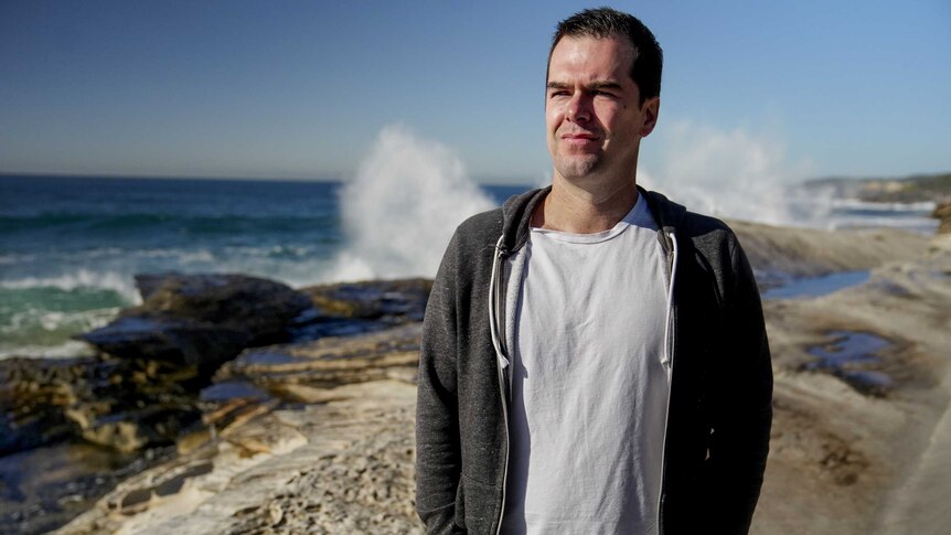 Man with black hair in white t-shirt and grey hoodie stands on beach with waves crashing on rocks behind him on a sunny day