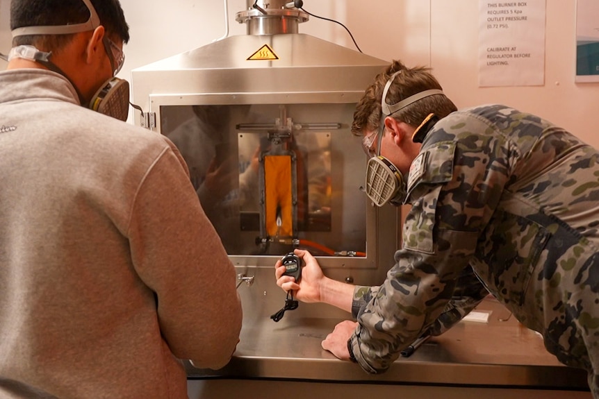 The backs of two men wearing masks looking at an oven-like machine. 