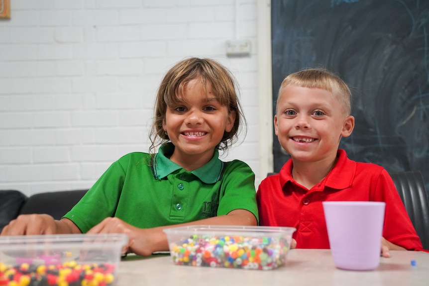 A boy wearing a red shirt and a girl wearing a green shirt smiling at the camera with a container of beads and a cup in front of