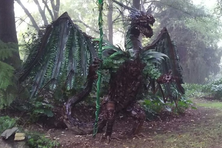 a 3 meter high dragon made from metal with spread wings  is nestled in a green garden on guard.