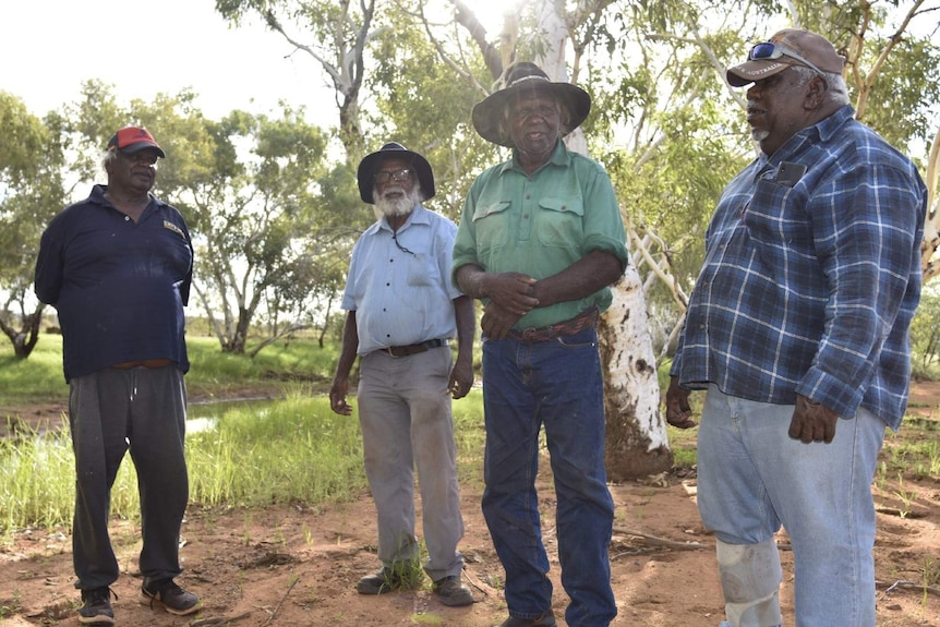 A group of four Indigenous men stand discussing something in the Central Australian outback.