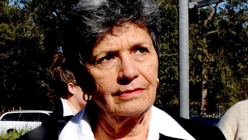 An elderly woman with short black hair, small round earrings, white, shirt and black jacket looks away from the camera. 